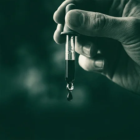 Fotografía en primer plano de un gotero de cristal transparente con aceite de cáñamo en posición vertical, sostenido entre el pulgar y el índice de una mano, con una gota de líquido justo formándose o cayendo desde la punta.