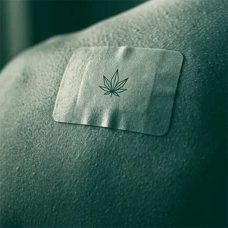 Fotografía en primer plano de un parche transdérmico rectangular ya aplicado sobre la piel (zona del hombro o gemelo), sin ninguna mano en el encuadre. Sobre la superficie del propio parche, un pequeño pictograma de hoja de cannabis impreso o grabado en el material, estilo geométrico y minimalista, líneas rectas y limpias.